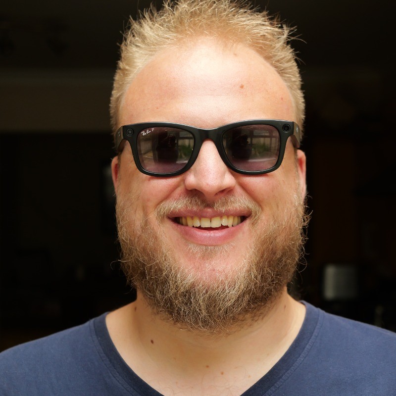 A happy person with a blonde beard and sunglasses is smiling broadly in a close-up indoor shot, with a dark background.