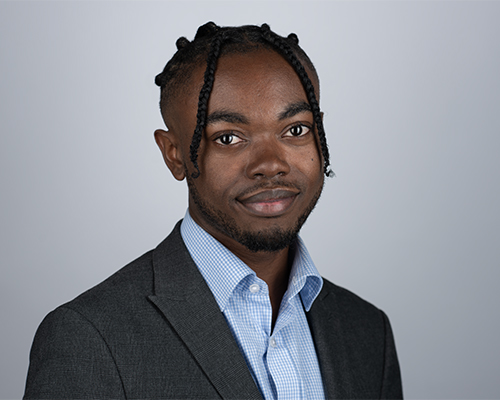 A professional-looking person with braided hair is smiling faintly while wearing a suit jacket in a studio with a grey backdrop.