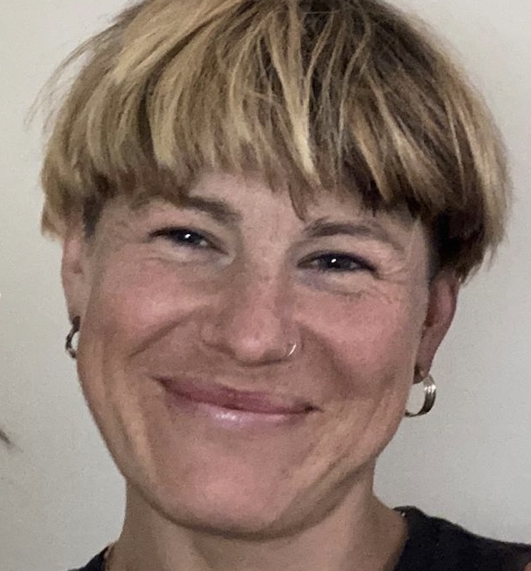 A pleasantly smiling person with short blonde hair and a nose ring is looking at the camera in an indoor, neutral-toned close-up.