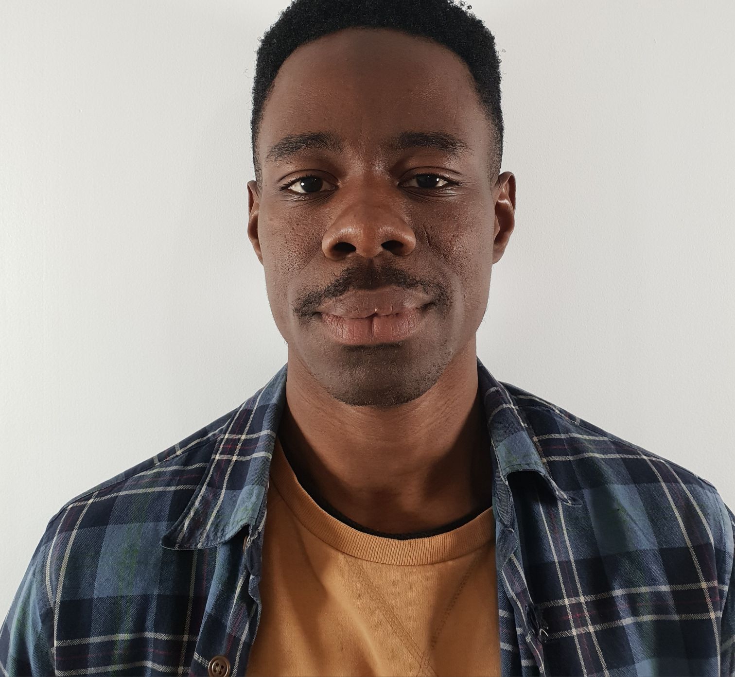 A calm person with a slight moustache is posing for a close-up portrait against a plain white wall.