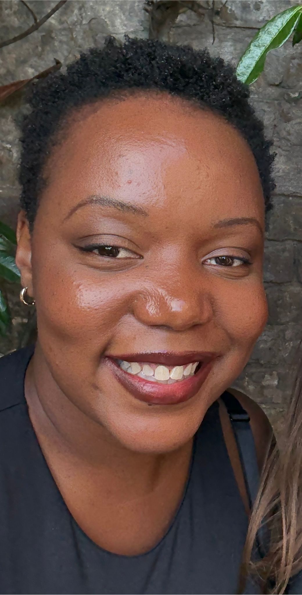 A joyful person with short, dark curly hair is smiling widely in a close-up outdoor photo against a brick wall backdrop.