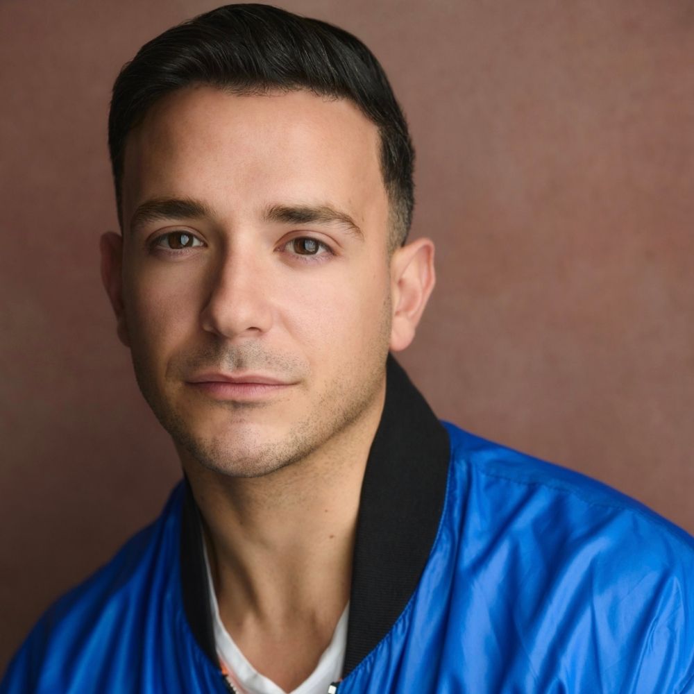 A contemplative person is posing in a bright blue jacket in a close-up indoor shot against a warm, brown background.